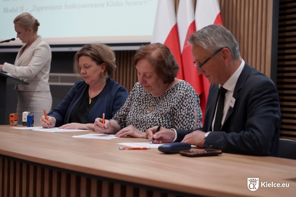 Trzy osoby siedzą przy stole i podpisują dokumenty. Patrząc od lewej strony to dwie kobiety i jeden mężczyzna.