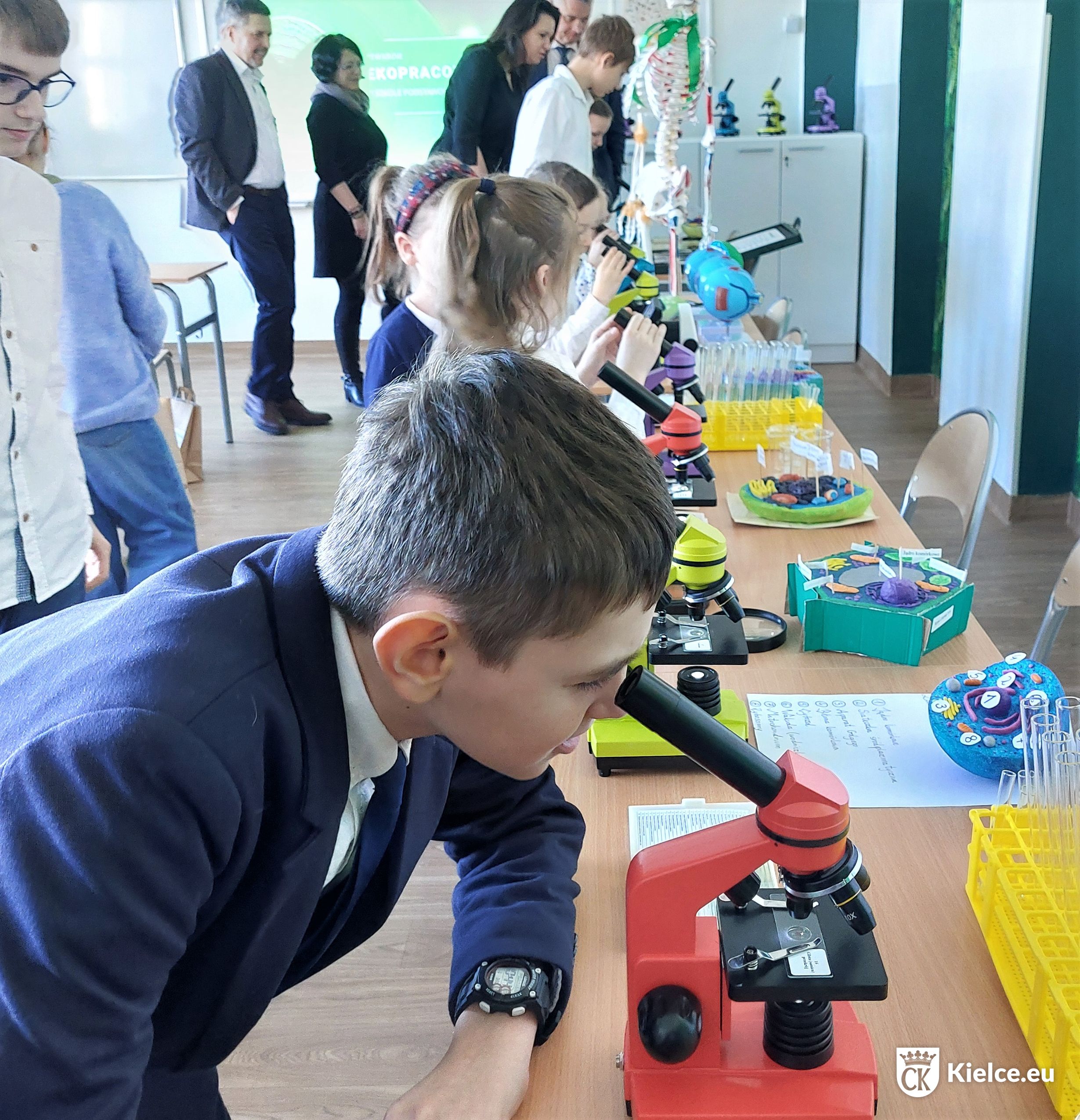 Na pierwszym planie chłopiec oglądający preparat przez mikroskop, w tle inne dzieci i dorośli oglądający nowy sprzęt w ekopracowni