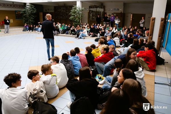 Duża grupa młodzieży siedzi na szkolnym holu. Na środku stoi mężczyzna z mikrofonem.