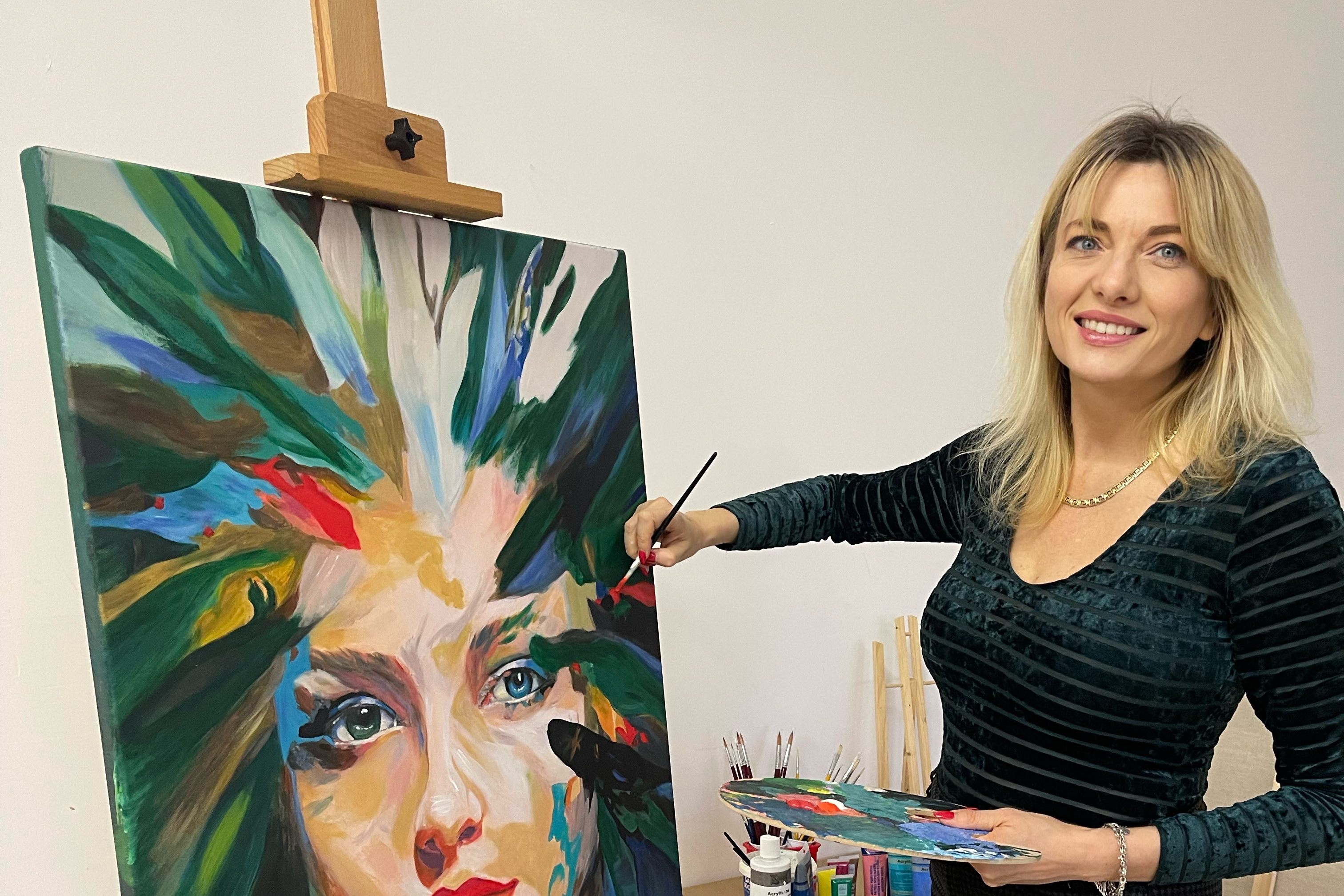 Kobieta ubrana na czarno maluje obraz. W lewej ręce trzyma paletę, w prawej pędzel. Na obrazie twarz kobiety, która zamiast włosów ma namalowane kolorowe promienie.