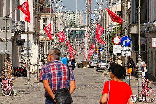 Na pierwszym planie odwrócony tyłem starszy mężczyzna w koszuli w kolorową kratkę i kobieta i czerwonej bluzce na ul. Sienkiewicza. Na drugim planie ulica i czerwone flagi z białym napisem "Budzenie Sienkiewki", zawieszone na latarniach miejskich.