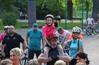 Kilku rowerzystów. Mężczyzna trzyma na ramionach kilkuletnia dziewczynkę, która pokazuje kciuk w górę.