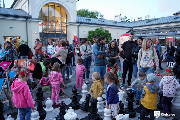 Grupa dzieci gra w wielkoformatowe szachy na dziedzińcu Muzeum Zabawek i Zabawy. Obok stoi grupa osób dorosłych.