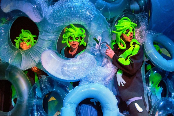 Aktorzy ubrani w zielone peruki imitujące włosy i czarne stroje stoją pomiędzy konstrukcją z dmuchanych kół ratunkowych.