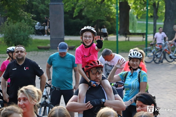 Mężczyzna w kasku rowerowym trzyma na ramionach dziewczynkę w różowej koszulce i kasku. Obok stoi kilka osób w strojach rowerowych.