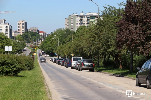 Ulica Bohaterów Warszawy, po prawej stronie zaparkowane samochody, drzewa, na drugim planie budynki. po lewej stronie jezdnia południowa, pas zieleni.