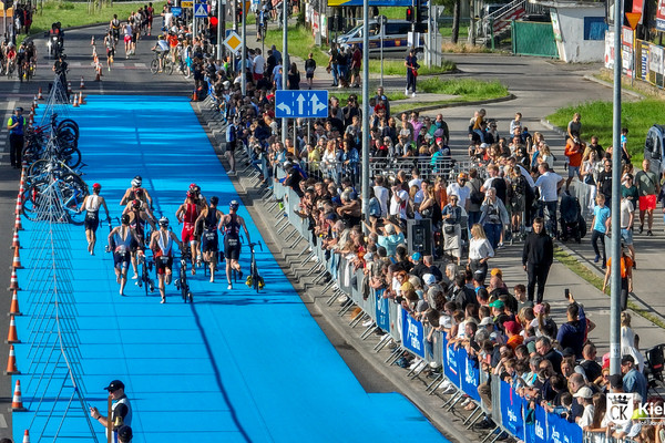 Zdjęcie z lotu ptaka, po prawej stronie kilkaset osób dopinguje zawodników, po środku grupa triathlonistów prowadzi rowery w strefie zmian.