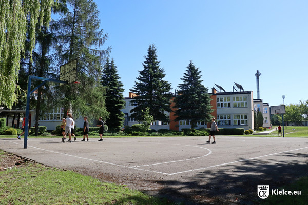 Kilku młodych ludzi gra w piłkę na asfaltowym boisku. Na drugim planie drzewa i budynki SP 28