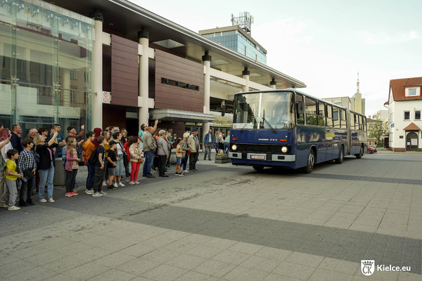Niebieski Ikarus stoi przed budynkiem parkingu wielopoziomowego. Po lewej stronie grupa osób czeka na autobus.