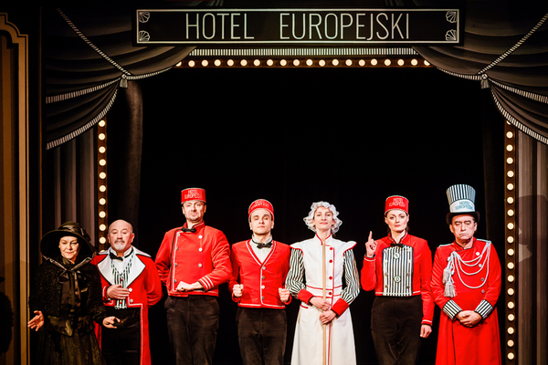 Grupa aktorów w czerwono-czarnych strojach pracowników hotelu stoi na scenie.