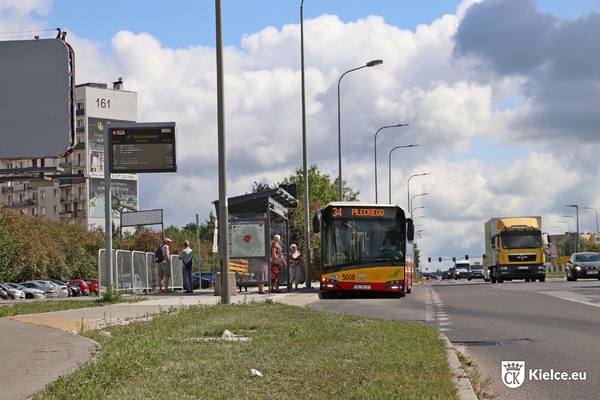 Przystanek przy ul. Warszawskiej i pasażerowie, na przystanku stoi autobus miejski, po prawej stronie kilka samochodów jedzie ulicą. Po lewej stronie bloki osiedla Uroczysko.