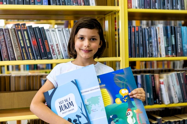 Dziewczynka z zestawem edukacyjnym w dłoniach, stoi na tle regału z książkami.