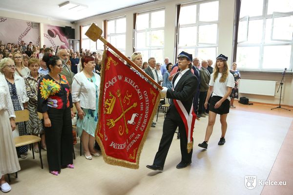 Młody mężczyzna niesie nowy sztandar szkoły, za nim idzie młoda kobieta, na drugim planie stoi kilkadziesiąt osób