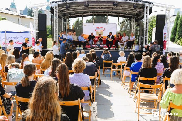 Na pierwszym planie, tyłem do fotografa siedzi publiczność, na drugim planie na scenie siedzi dwanaście osób