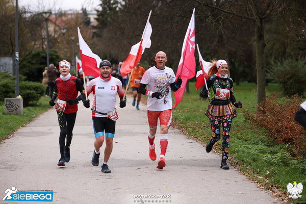 Czworo biegaczy, troje trzyma biało-czerwone flagi, na drugim planie grupa biegaczy